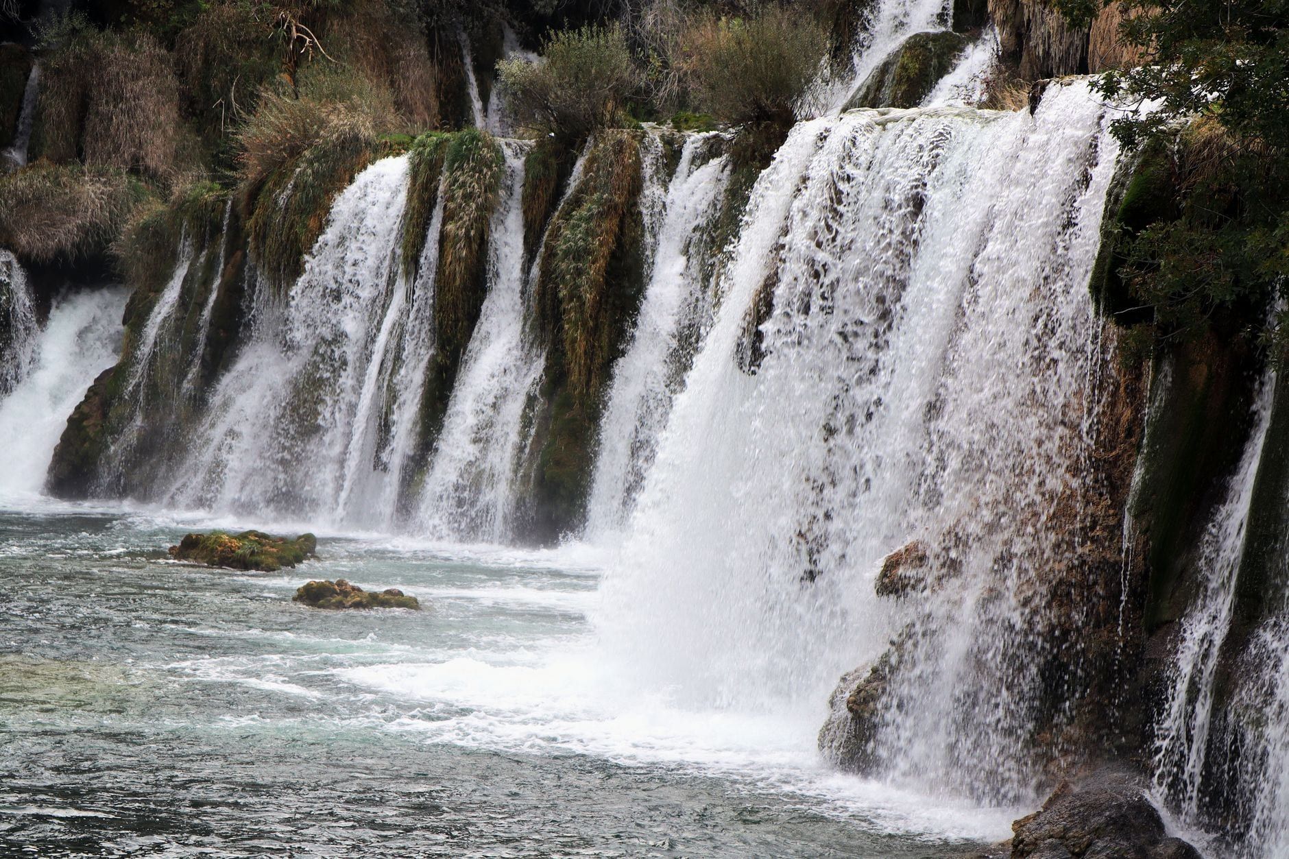 Wasser kaskadiert über felsige, moosbewachsene Stufen in ein Flussbecken im Krka-Nationalpark.