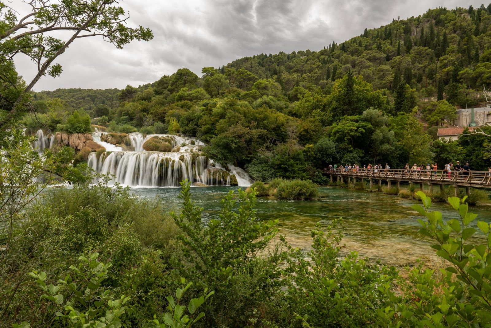 Des visiteurs marchent sur une passerelle en bois traversant le bassin clair de la rivière devant la cascade de Skradinski buk dans le parc national de Krka.