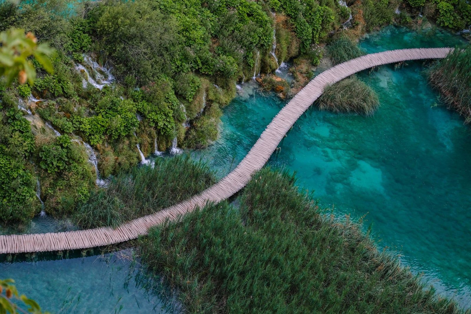 A wooden boardwalk path winds across turquoise water alongside lush green vegetation and small waterfalls in Plitvice Lakes National Park.