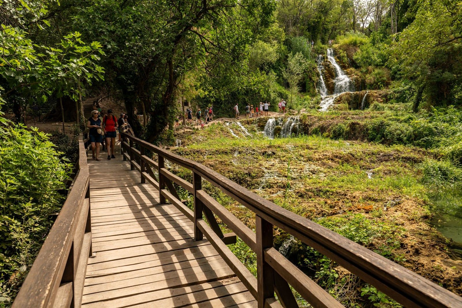 Visitantes caminando por una pasarela de madera rodeada de vegetación exuberante y cascadas en el Parque Nacional de Krka.